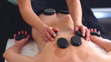 Terapist sıcak taş masajı yapıyor, müşterilerin sırtına pürüzsüz siyah taşlar sürüyor. Rahatlatıcı sağlık tedavisine yakın çekim. Spa, terapi, rahatlama ve bütünsel iyileşme kavramları için ideal..