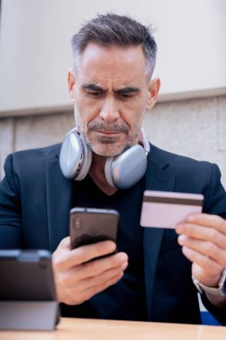Kulaklıklı, akıllı telefon kullanırken gülümseyen ve dışarıda kredi kartı tutan kendine güvenen beyaz bir adam. Çevrimiçi alışveriş kavramı, dijital ödeme, modern yaşam tarzı ve finansal teknoloji. 