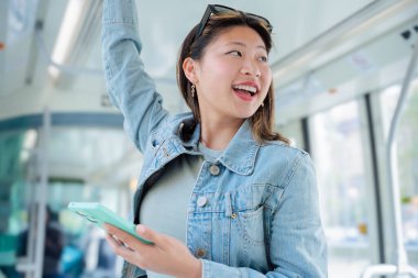 Mutlu Çinli kadın bir elinde tramvay korkuluğuna tutunurken diğer elinde akıllı telefon kullanıyor. Kentsel toplu taşımada gülümsüyor. Yaşam tarzı, teknoloji, seyahat ve işe gidiş gelişler için mükemmel..