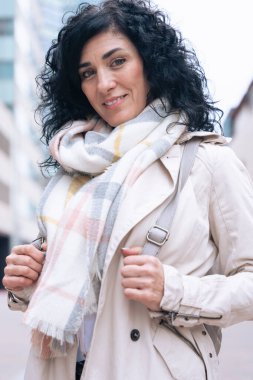 Yakından kendine güvenen, kıvırcık siyah saçlı, atkı ve palto giyen, şehir ortamında dışarıda duran beyaz bir kadın. Modern yaşam tarzı, şehir modası ve kendinden emin ifade serin bir günde. 