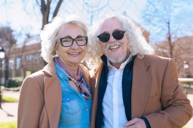 Kafkasyalı yaşlı bir çift dışarıda fotoğraf çektirmek için kameraya bakıp gülümsüyor. Gözlükleri ve kahverengi ceketleri ile birlikte güneşli bir günün tadını çıkarıyorlar.. 