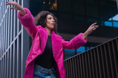 Pembe palto giyen kıvırcık saçlı zarif dansçı şehir ortamında hareketini ve enerjisini ifade ediyor. Modern moda, çağdaş dans ve sanatsal performans şehrin arka planına karşı..