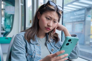 Genç Çinli kadın toplu taşıma aracının penceresinde oturmuş endişeli bir ifadeyle akıllı telefonunu okuyor. Şehir içi bir ortamda günlük kıyafetler ve güneş gözlüğü takıyor.. 