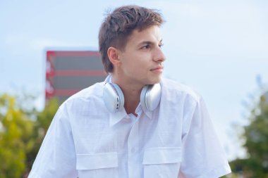 Boynuna beyaz gömlek ve kulaklık takmış genç beyaz bir adam düşünceli bir şekilde başka tarafa bakıyor. Modern yaşam tarzı, moda ve müzik atmosferi güneşli açık şehir atmosferinde.
