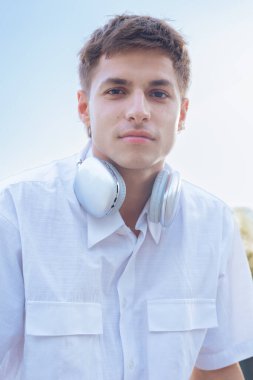 Boynuna beyaz gömlek ve kulaklık takmış beyaz bir adam doğrudan kameraya bakıyor. Açık hava portresi, sakin bir ifade ve doğal gün ışığı altında modern bir gündelik tarz.