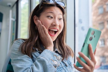 Trenin penceresinde oturan genç Çinli kadın akıllı telefona bakarken şaşkınlıkla gülümsüyor. Günlük kıyafet ve neşeli ifade seyahat ederken eğlenceli ve spontane bir dijital anı yakalıyor. 