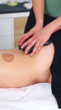 Terapist sıcak taş masajı yapıyor, müşterilerin sırtına pürüzsüz siyah taşlar sürüyor. Rahatlatıcı sağlık tedavisine yakın çekim. Spa, terapi, rahatlama ve bütünsel iyileşme kavramları için ideal.