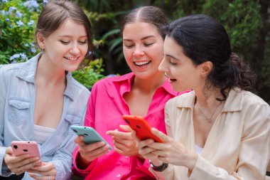 Üç beyaz kadın yeşil bir parkta ahşap bir bankta oturuyor, akıllı telefonlar kullanırken gülümsüyorlar. Mutlu ve meşgul görünüyorlar, modern cihazlar ve sosyal medya uygulamalarıyla açık havada eğleniyorlar.
