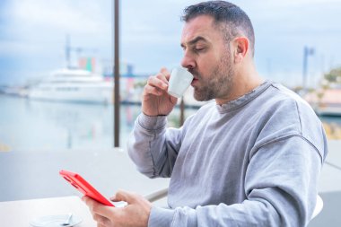 Kısa siyah saçlı ve sakallı bir adam akıllı telefonuyla konuşurken kahve içer. Günlük gri bir kazak giyer ve açık havada suyun yanında durur. Modern şehir ortamına odaklanmış gibi görünür.