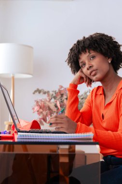 Afro-Amerikalı kadın evde oturmuş laptopuyla uğraşırken düşünüyor. O bir kalem tutar ve düşünceli bir şekilde uzağa bakar. Uzaktan çalışma, eğitim, odaklanma ve dijital yaşam tarzı kavramı. Dikey