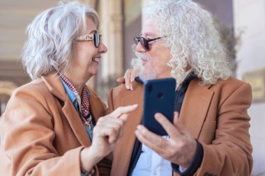 Birlikte akıllı telefon kullanırken gülümseyen Kafkasyalı son sınıf çifti. Şık ceketler ve gözlük takıyorlar, dışarıda mutlu bir anın tadını çıkarıyorlar. Aşk kavramı, bağlantı ve çağdaş yaşam tarzı.. 