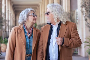 Güneş gözlüğü takan, kahverengi ceketli şık bir çift dışarıda yürüyor. Kendilerinden emin ve rahatlar. Mimari unsurları olan bir şehir ortamında olgun moda ve arkadaşlıklar sergiliyorlar.. 