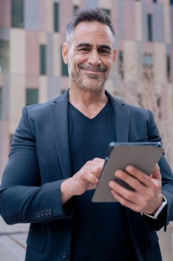 Kendine güveni tam bir işadamı modern şehir ortamında tablet kullanırken gülümsüyor. Siyah bir takım elbise ve şık bir elbise giyiyor. Başarı kavramı, teknoloji, dijital yaşam tarzı ve üretkenlik. Dikey 