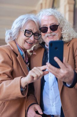 Birlikte akıllı telefon kullanırken gülümseyen Kafkasyalı son sınıf çifti. Şık ceketler ve gözlük takıyorlar, dışarıda mutlu bir anın tadını çıkarıyorlar. Aşk kavramı, bağlantı ve çağdaş yaşam tarzı.. 