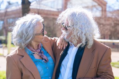 Güneşli bir parkta dikilen, gülümseyen ve birlikte poz veren beyaz yaşlı bir çift. Kadın kolunu erkeğin omzuna koyar. Bu arada onlar da şehrin tadını çıkarırlar.. 