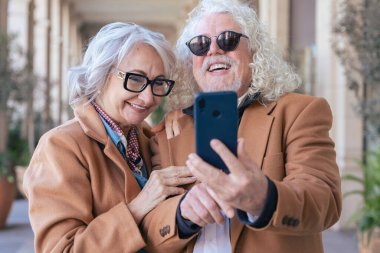 Kafkasyalı mutlu çift akıllı telefonuyla selfie çekiyor. Güneş gözlüğü ve ceket giyerek, parlak bir şekilde gülümserler. Açık havada aşk, neşe, bağlantı ve modern yaşam tarzı kavramı. 