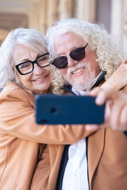 Kafkasyalı mutlu çift akıllı telefonuyla selfie çekiyor. Güneş gözlüğü ve ceket giyerek, parlak bir şekilde gülümserler. Açık havada aşk, neşe, bağlantı ve modern yaşam tarzı kavramı. 
