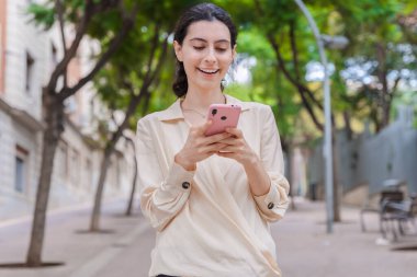 Ağaçlarla kaplı bir şehir sokağında akıllı telefonunu kullanırken gülümseyen beyaz bir kadın. Şehirde keyifli bir doğa yürüyüşü sırasında rahatlamış ve cihazıyla meşgul görünüyor..