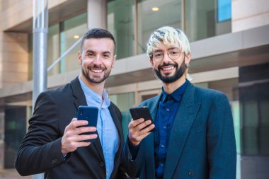 İki neşeli, kendinden emin iş adamının portresi, gülümseyen ve akıllı telefonlar tutan, resmi kıyafetler giyen, modern ofis binalarının yanında duran, kameraya olumlu bakan 