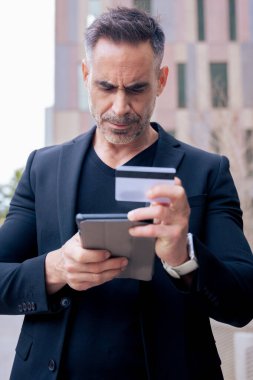 Kulaklıklı, kendine güvenen beyaz adam tablet kullanırken ve dışarıda kredi kartı tutarken ciddiydi. Çevrimiçi alışveriş, dijital ödeme, modern yaşam tarzı ve finansal teknoloji kavramı. Dikey