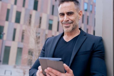 Kendine güveni tam bir işadamı modern şehir ortamında tablet kullanırken gülümsüyor. Siyah bir takım elbise ve şık bir elbise giyiyor. Başarı kavramı, teknoloji, dijital yaşam tarzı ve üretkenlik. Boşluğu kopyala