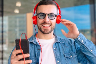Gülümseyen beyaz adam elinde akıllı bir telefonla kırmızı kulaklıkla müzik dinlemekten hoşlanıyor. Kot gömlek giymiş, arka planda modern cam bina olan açık havada duran, parlak ve sıradan bir an.. 