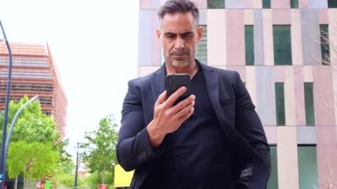 Siyah kıyafetli, kendine güvenen beyaz adam kameraya doğru ciddi bir yüzle akıllı telefon tutuyor. Modern mimariyle kentsel bir çevrede iş, teknoloji ve iletişim kavramı. 