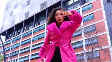 Pembe palto giyen kıvırcık saçlı zarif dansçı şehir ortamında hareketini ve enerjisini ifade ediyor. Modern moda, çağdaş dans ve sanatsal performans şehrin arka planına karşı..