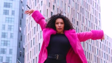 Pembe palto giyen kıvırcık saçlı zarif dansçı şehir ortamında hareketini ve enerjisini ifade ediyor. Modern moda, çağdaş dans ve sanatsal performans şehrin arka planına karşı..