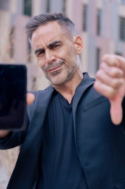 Siyah kıyafetli bir iş adamı akıllı telefonunu gösteriyor ve kameraya bakarak modern teknolojinin başarısını ve yok oluşunu öneren kentsel bir arka planda kendine güvenen bir şekilde başparmağıyla onaylıyor. 