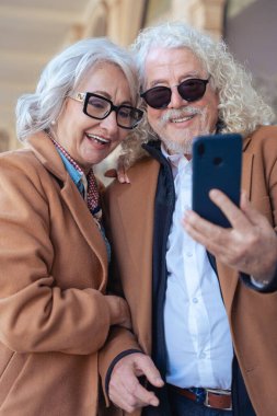 Birlikte akıllı telefon kullanırken gülümseyen Kafkasyalı son sınıf çifti. Şık ceketler ve gözlük takıyorlar, dışarıda mutlu bir anın tadını çıkarıyorlar. Aşk kavramı, bağlantı ve çağdaş yaşam tarzı.. 