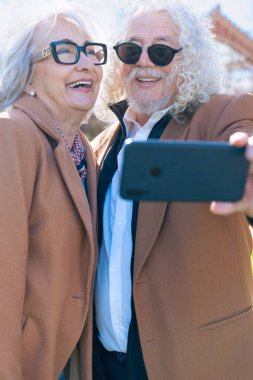 Mutlu Kafkasyalı yaşlı bir çift bankta oturmuş dışarıda selfie çekiyorlar. Kahverengi ceket ve gözlük takarlar, güneşli bir günün tadını çıkarırlar ve o anı akıllı bir telefonla yakalarlar.. 