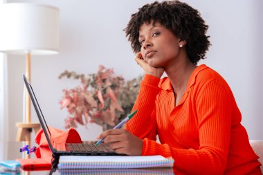 Afro-Amerikalı kadın evde oturmuş laptopuyla uğraşırken düşünüyor. O bir kalem tutar ve düşünceli bir şekilde uzağa bakar. Uzak çalışma, eğitim, odaklanma ve dijital yaşam tarzı kavramı. 