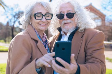 Kafkasyalı yaşlı bir çift sosyal medyayı kontrol etmek için akıllı telefon uygulaması kullanıyor. Ekranı paylaşırken gülümserler, güneşli bir günün keyfini çıkarırlar günlük kahverengi ceketler ve gözlüklerle.. 