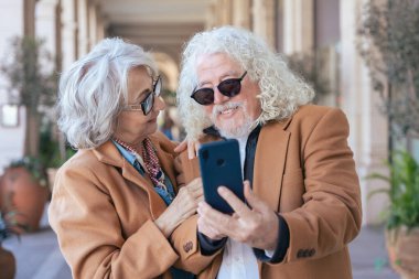 Birlikte akıllı telefon kullanırken gülümseyen Kafkasyalı son sınıf çifti. Şık ceketler ve gözlük takıyorlar, dışarıda mutlu bir anın tadını çıkarıyorlar. Aşk kavramı, bağlantı ve çağdaş yaşam tarzı.. 