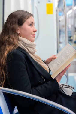  Siyah paltolu ve bej eşarplı genç bir kadın modern bir metro treninde oturmuş, odaklanmış bir kitap okuyor. Şehirdeki seyahati sırasında sakin bir anın tadını çıkarıyor..