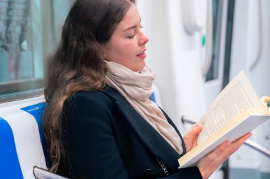  Siyah paltolu ve bej eşarplı genç bir kadın modern bir metro treninde oturmuş, odaklanmış bir kitap okuyor. Şehirdeki seyahati sırasında sakin bir anın tadını çıkarıyor..