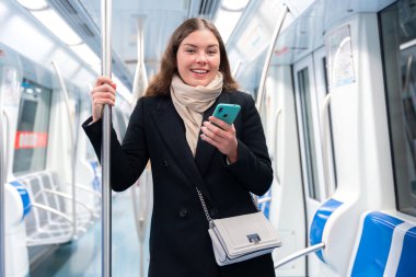 Siyah paltolu ve bej eşarplı neşeli genç bir kadın modern bir metro treninin içinde elinde akıllı bir telefon ve destek için metal bir direği tutuyor. Seyahat ederken ışıl ışıl gülümsüyor.. 
