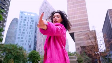 Pembe palto giyen kıvırcık saçlı zarif dansçı şehir ortamında hareketini ve enerjisini ifade ediyor. Modern moda, çağdaş dans ve sanatsal performans şehrin arka planına karşı..
