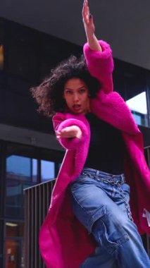 Pembe palto giyen kıvırcık saçlı zarif dansçı şehir ortamında hareketini ve enerjisini ifade ediyor. Modern moda, çağdaş dans ve sanatsal performans şehrin arka planına karşı..