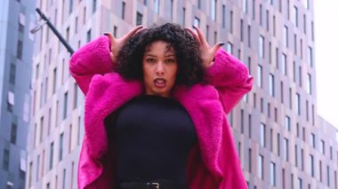 Pembe palto giyen kıvırcık saçlı zarif dansçı şehir ortamında hareketini ve enerjisini ifade ediyor. Modern moda, çağdaş dans ve sanatsal performans şehrin arka planına karşı..