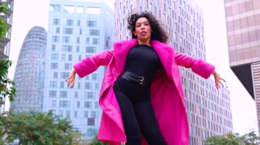 Pembe palto giyen kıvırcık saçlı zarif dansçı şehir ortamında hareketini ve enerjisini ifade ediyor. Modern moda, çağdaş dans ve sanatsal performans şehrin arka planına karşı..