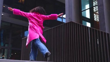 Pembe palto giyen kıvırcık saçlı zarif dansçı şehir ortamında hareketini ve enerjisini ifade ediyor. Modern moda, çağdaş dans ve sanatsal performans şehrin arka planına karşı..