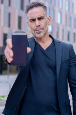 Siyah kıyafetli, kendine güvenen beyaz adam kameraya doğru ciddi bir yüzle akıllı telefon tutuyor. Modern mimariyle kentsel bir çevrede iş, teknoloji ve iletişim kavramı. 