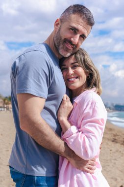 Tatil sırasında sahilde kucaklaşan gülümseyen beyaz bir çiftin dikey portresi. Deniz kenarında sakin ve sevecen bir an. Mavi gökyüzü, okyanus dalgaları ve arkalarında kumlu bir sahil.. 