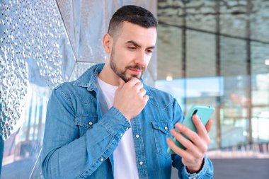 Kot gömlek giyen genç bir adam, yansıtıcı bir cam duvarın yanında dikiliyor, akıllı telefonuna özenle bakıyor, iletişimi ve modern hayatı sembolize ediyor.. 