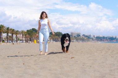 Genç bir kadın, güneşli bir kumsalda, palmiye ağaçlarıyla Bernese Mountain Dog 'la yürüyor. Mavi gökyüzünün altında rahatlatıcı bir an, deniz kenarında yürümek için mükemmel bir gün, sıradan ve doğal bir sahne.. 