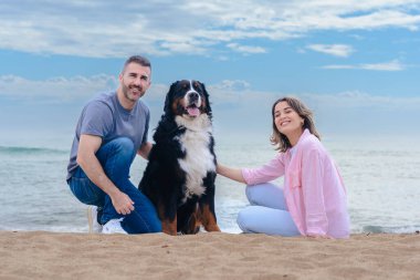 Tatil sırasında kumsalda büyük bir köpekle gülümseyen beyaz bir çiftin portresi. Deniz kenarında rahat ve keyifli bir an. Üçü de aydınlık bir günde kameraya bakıyor.. 