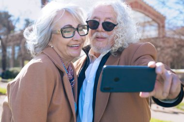 Mutlu Kafkasyalı yaşlı bir çift bankta oturmuş dışarıda selfie çekiyorlar. Kahverengi ceket ve gözlük takarlar, güneşli bir günün tadını çıkarırlar ve o anı akıllı bir telefonla yakalarlar.. 