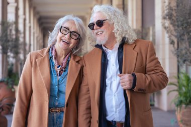 Dışarıda yürüyen, güneş gözlüğü ve kahverengi ceketli gülümseyen son sınıf çifti. Mimari geçmişi ve bitkileri olan şık bir şehir ortamında zarafet, neşe ve arkadaşlık gösterirler.. 